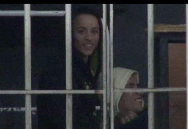 2 bandidos sorrindo atrás de grades, em delegacia. Um está com capuz branco, o outro sem