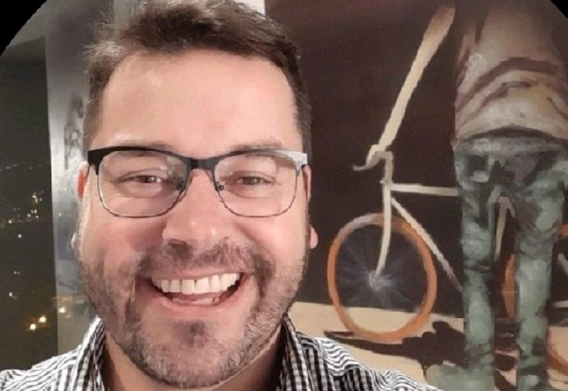 Advogado José Rodrigo Sade sorrindo em frente a pintura que retrata uma pessoa segurando uma bicicleta (Reprodução/LinkedIn)