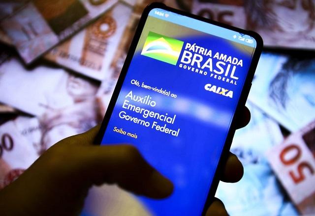 Pessoa segura celular com aplicativo da Caixa aberto, em frente a cédulas de reais em diferentes valores (Marcelo Camargo/Agência Brasil)