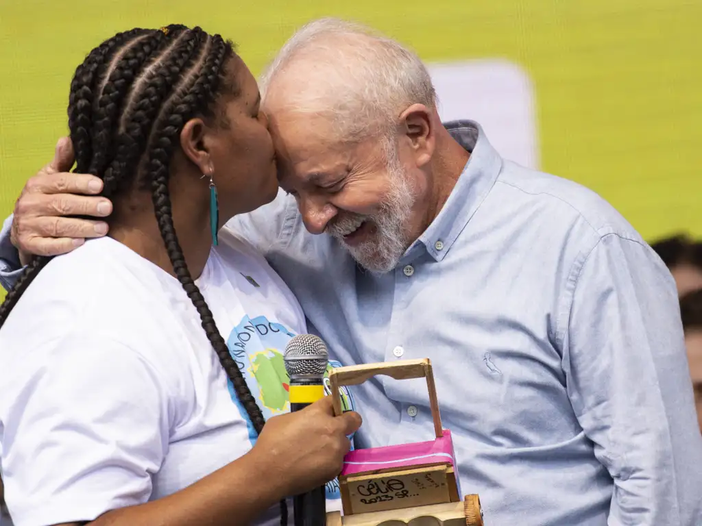 Mulher negra com cabelos trançados beija a testa de Lula em evento de comemoração com catadores.