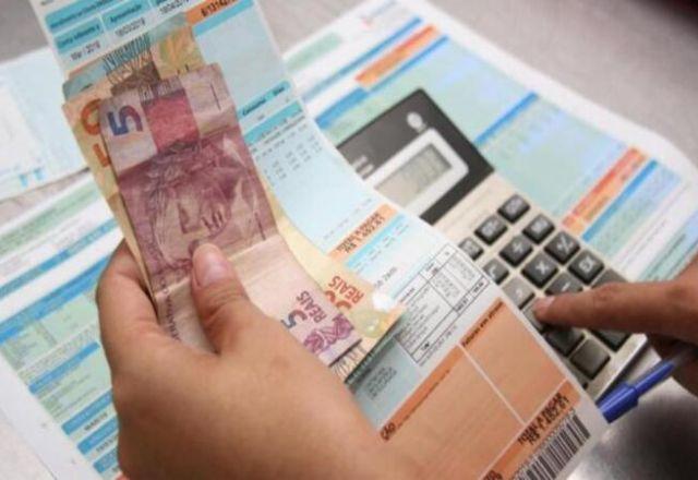 Pessoa mexe em calculadora enquanto segura conta e cédulas de real (Reprodução/Agência Brasil)