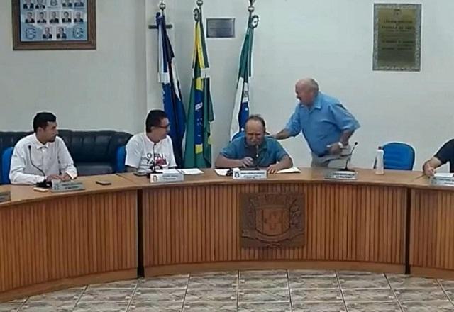 Bancada de câmara de vereadores, com um homem idoso de pé cumprimentando outros três sentados