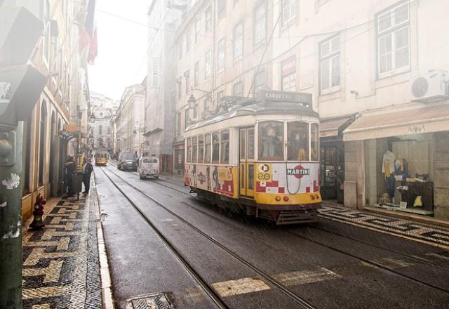 Bonde em uma rua de paralelepípedo com uma vitrine de roupas ao lado e outros automóveis ao fundo