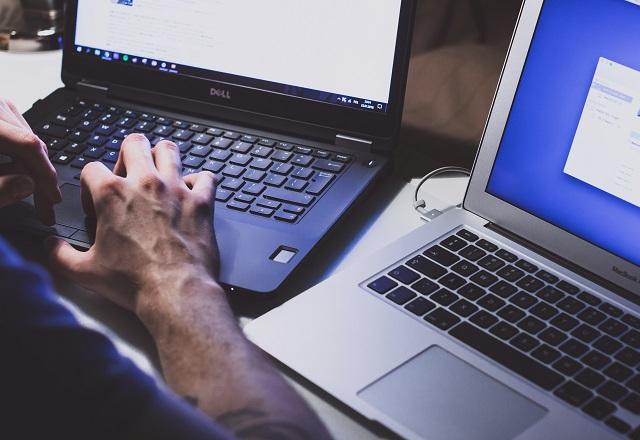 Braço de homem trabalhando com dois notebooks em mesa, os dois notebooks com as telas iluminadas e abertas