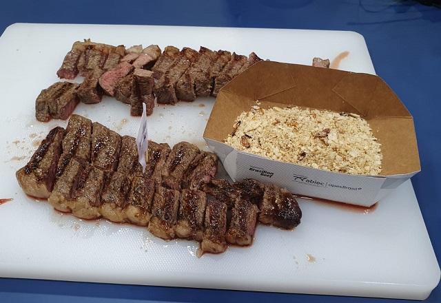 Carne cortada em pedaços ao lado de recipiente com farofa, sobre bandeja