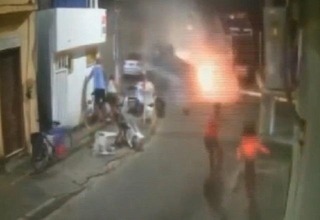 Carro após capotar, pegando fogo no meio de rua, com pessoas ao redor correndo em desespero
