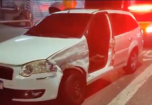 Carro branco sem porta do lado do motorista, com avarias após acidente, e viatura policial com giroflex ligado atrás