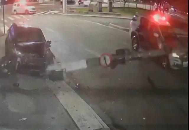 Carro colidido contra poste, com poste derrubado e caído sobre capô de viatura policial em rua durante a noite