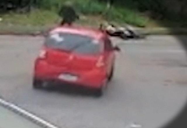Carro vermelho acelerando para cima de homem e moto, no meio da rua, durante o dia