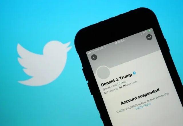 Celular, com o aplicativo do Twitter aberto, mostra que o perfil de Donald Trump está bloqueado. Ao fundo, o logo da rede social