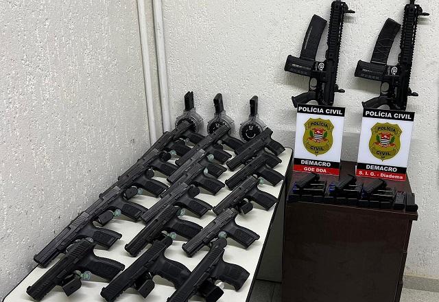Dezenas de armas do tipo pistola dispostas sobre mesa, com dois fuzis em pé ao fundo