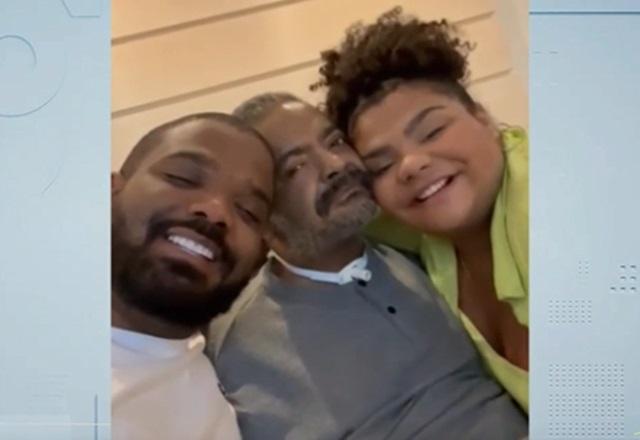 Dois homens negros e uma mulher negra, lado a lado, abraçados e sorrindo
