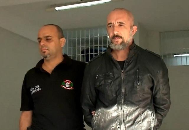 Dois homens, um de barba grisalha e vestindo jaqueta preta, sendo escoltado por policial com camiseta preta