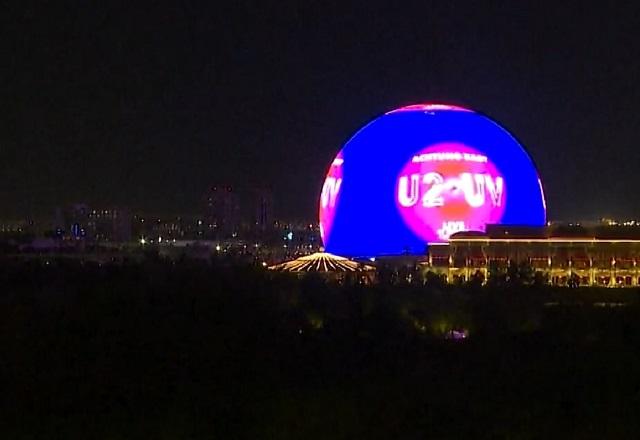 Enorme arena en formato de esfera, com fachada externa totalmente coberta por iluminação, no meio de prédios durante a noite
