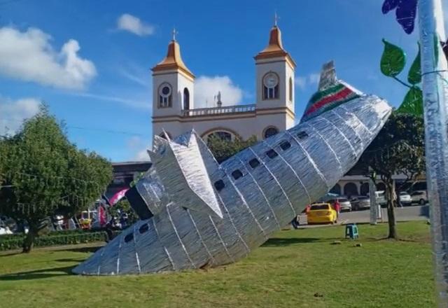 Estrutura de alumínio em formato de avião caído no solo, em praça, durante o dia