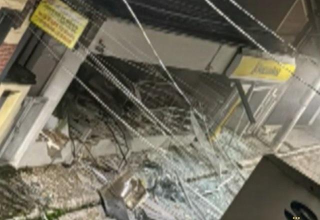 Fachada de agência bancária destruída após explosão, com vidro quebrado e ferro retorcido espalhado