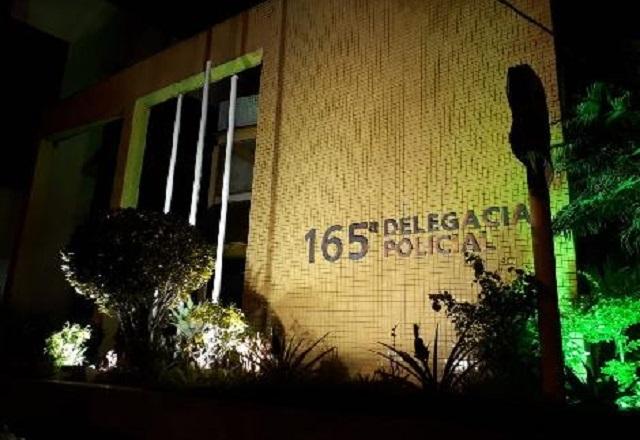 Fachada de delegacia, com nome do local inscrito, iluminado, durante a noite
