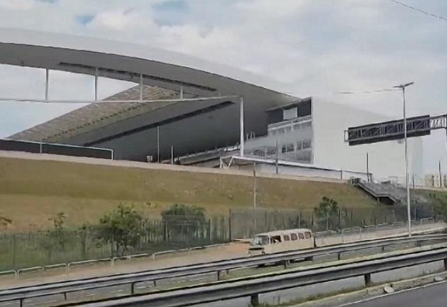 Fachada de estádio de futebol, mostrando canto e parte da arquibancada, em imagem vista de avenida, com placas e guard-rails no meio da via, durante o dia