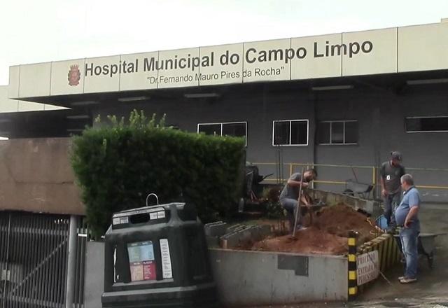 Fachada de hospital, durante o dia, com homens trabalhando em canteiro e lidando com terra