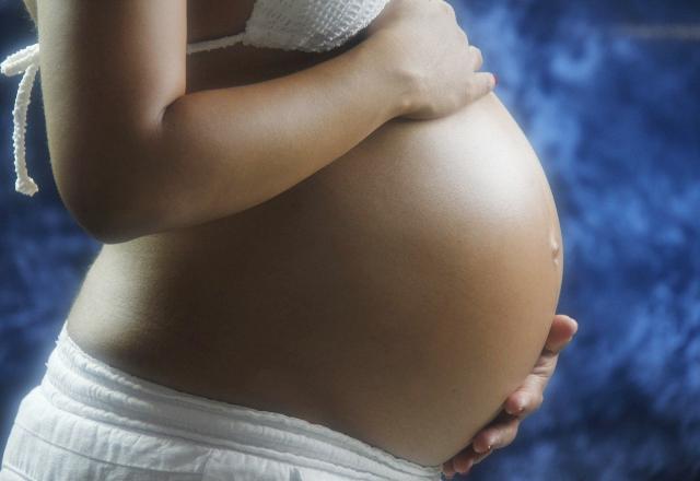 Foto da barriga de uma mulher grávida. Ela veste sutiã e shorts brancos. O fundo da imagem é azul