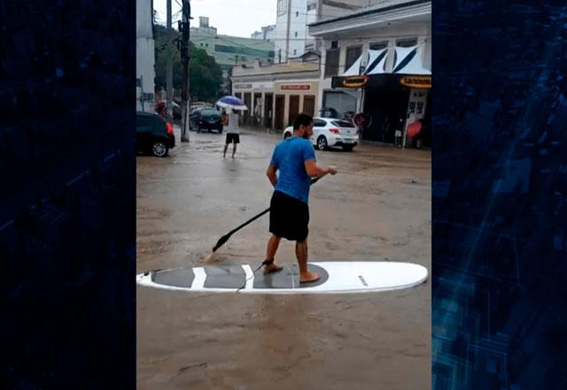 Foto de homem com camisa azul e shorts preto em cima de uma prancha, segurando um remo. Ao redor, lojas fechadas por causa da água da enchente