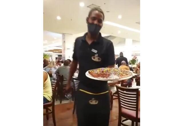 Garçom com uniforme preto, máscara cobrindo nariz e boca, segurando uma bandeja