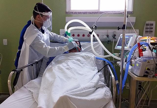 Profissional da saúde segura respirador mecânico sobre o rosto de um paciente deitado em cama, dentro de leito hospitalar (Thiago Conrado/Governo do Ceará)