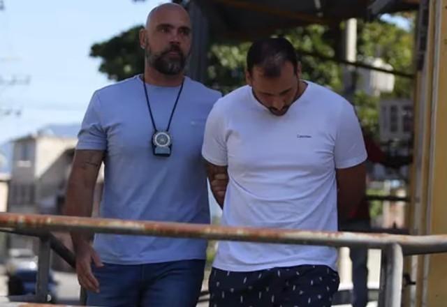 Homem algemado com camisa branca, sendo levado por policial de camisa azul