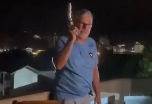 Homem branco, de cabelos grisalhos, com arma em punho disparando para o alto durante festa à noite