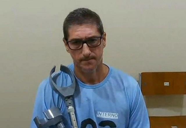 Homem branco, de camiseta azul de detento, com muleta amparada no peito, sentado durante depoimento