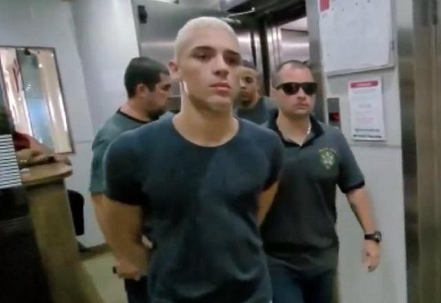 Homem branco, de camiseta cinza, com as mãos para trás sendo escoltado durante prisão