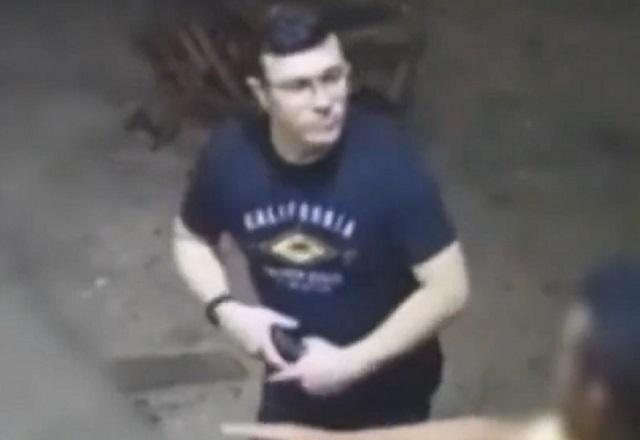 Homem branco, de camiseta preta e óculos, com arma nas mãos, aparentemente descontrolado