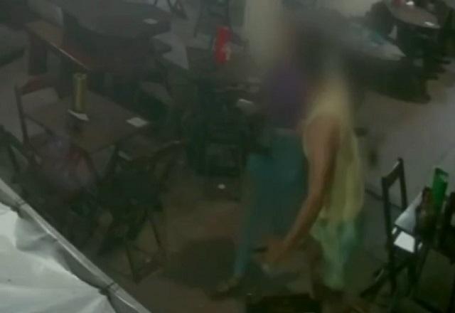 Homem branco, de camiseta regata amarela amarela, agredindo mulher de blusa roxa, entre mesas e cadeiras de bar