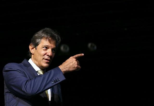 Homem branco, de terno preto, sob fundo escuro, apontando o dedo em direção à frente