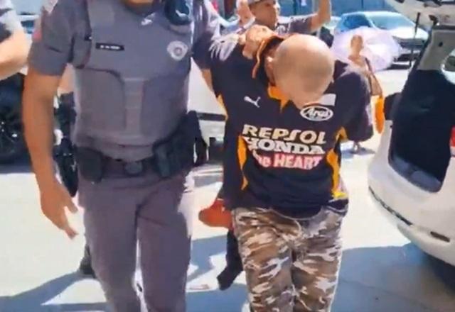 Homem branco e careca, algemado e sendo conduzido por policial durante prisão