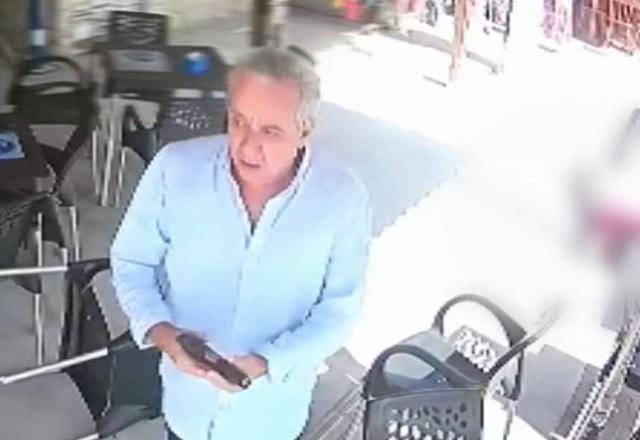 Homem branco idoso, de camisa branca, segurando arma após atirar em vítima, de camiseta vinho, caída no chão no fundo da imagem