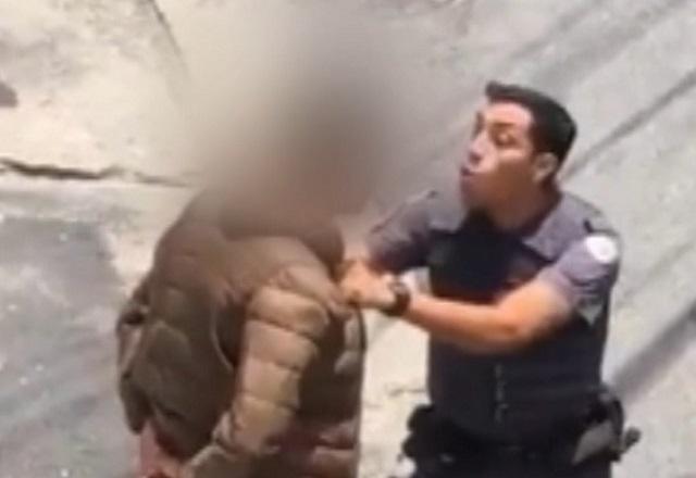 Homem branco policial de farda, segurando outro pelo colarinho da blusa, com rosto desfocado para nao identificação, durante abordagem policial violenta