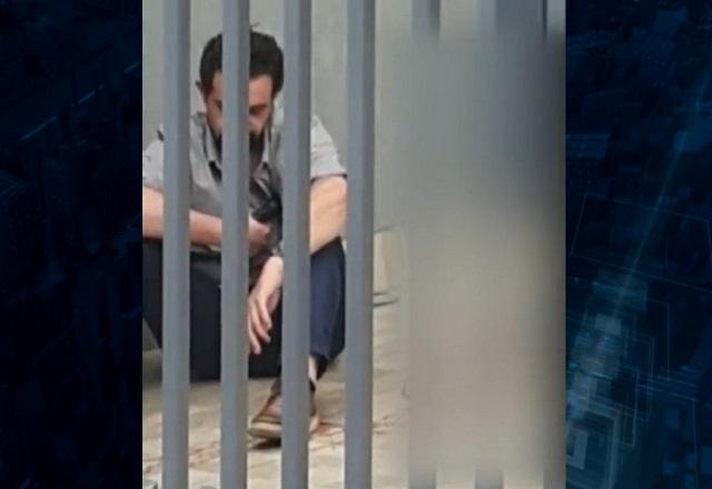Homem branco, sentado no chão atrás de portão, segurando arma