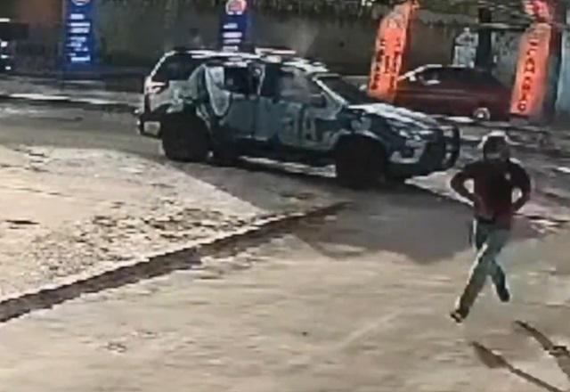 Homem correndo em rua tentando escapar de tiros disparados de viatura policial ao fundo