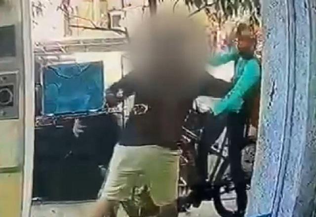 Homem de blusa escura dando soco em entregador, de camiseta verde e a bordo de bicicleta, em frente a porta de vidro de prédio