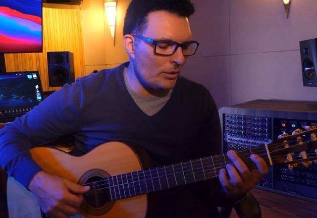 Homem de camisa preta, tocando violão, dentro de estúdio musical