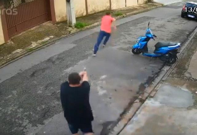 Homem de camiseta preta e armado aponta arma para homem correndo, de camiseta rosa, no meio da rua