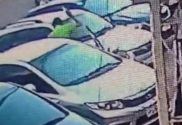 Homem de camiseta verde, com arma apontada para o alto, entre dois veículos estacionados