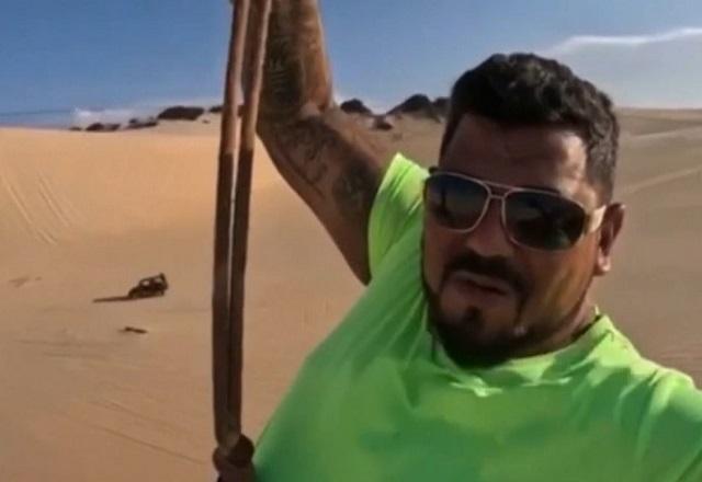 Homem de camiseta verde e óculos escuro segurando-se em corda durante descida de tirolesa em areias