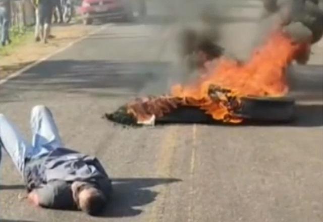 Homem deitado e amarrado em asfalto de rodovia, ao lado de barricada de fogo