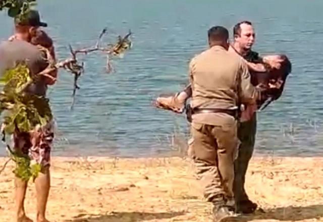 Homem fardado segurando mulher nos braços, durante o dia, em margem de rio durante operação de resgate