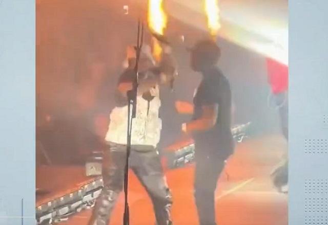 Homem negro cantando em palco, com microfone na mão, fazendo movimento antes de arremessar o item na direção da plateia