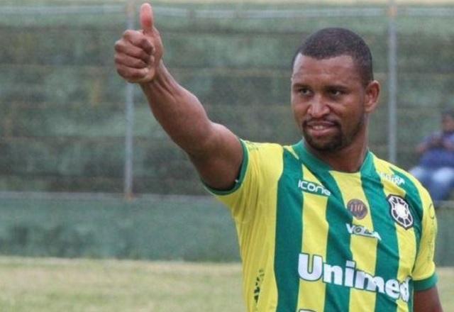 Homem negro, com uniforme de futebol verde e amarelo, com o braço levantado e fazendo sinal de joia