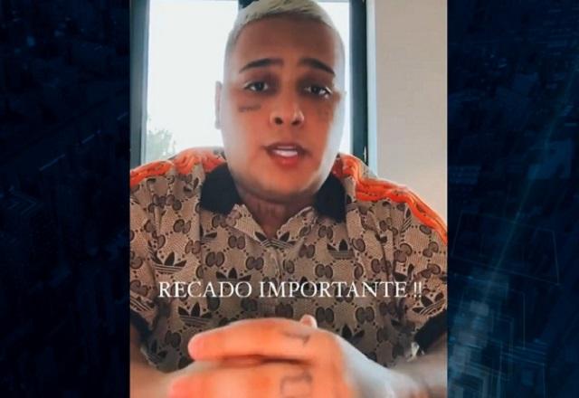 Homem negro, de cabelos tingidos de loiro e vestindo camisa estampada bege, com os dedos da mão cruzados enquanto olha para a câmera