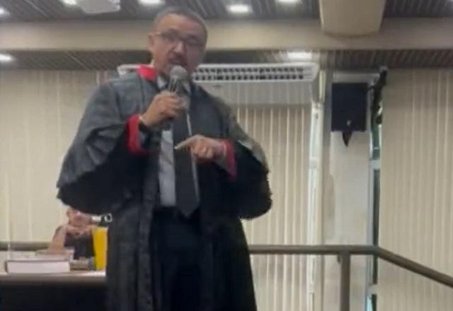 Homem negro de toga e óculos, segurando microfone,durante audiência em tribunal